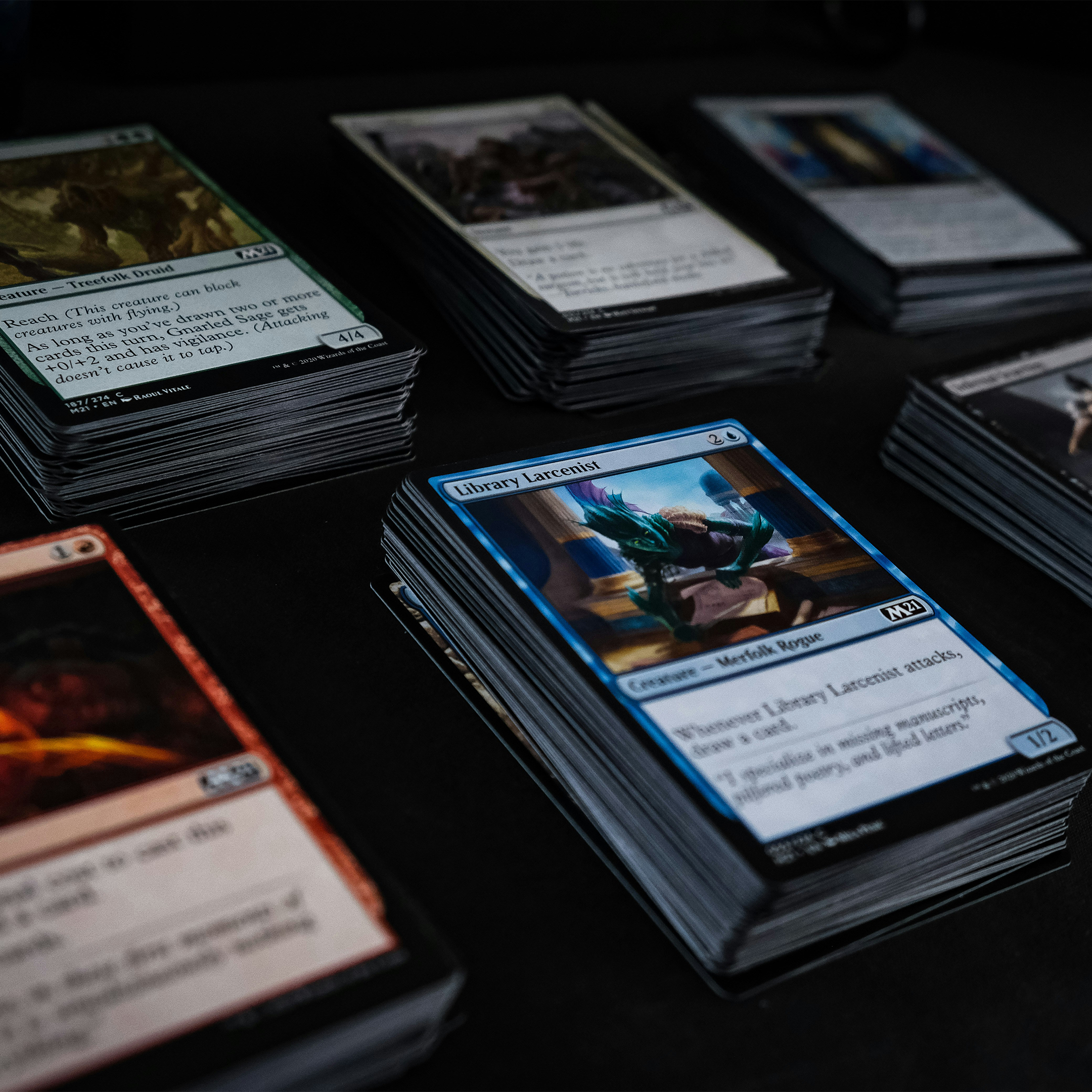A close-up, angled view of several Magic: The Gathering trading cards arranged on a dark surface. The central card is a blue Library Locus card, with other cards fanned out around it, showing various artwork and text.
